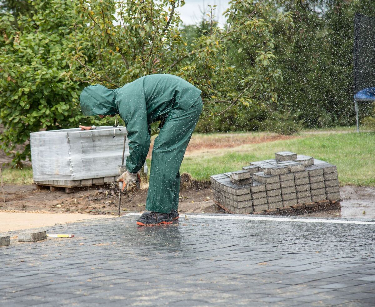 Een stratenmaker in regenpak legt grijze klinkers op een nieuwe oprit